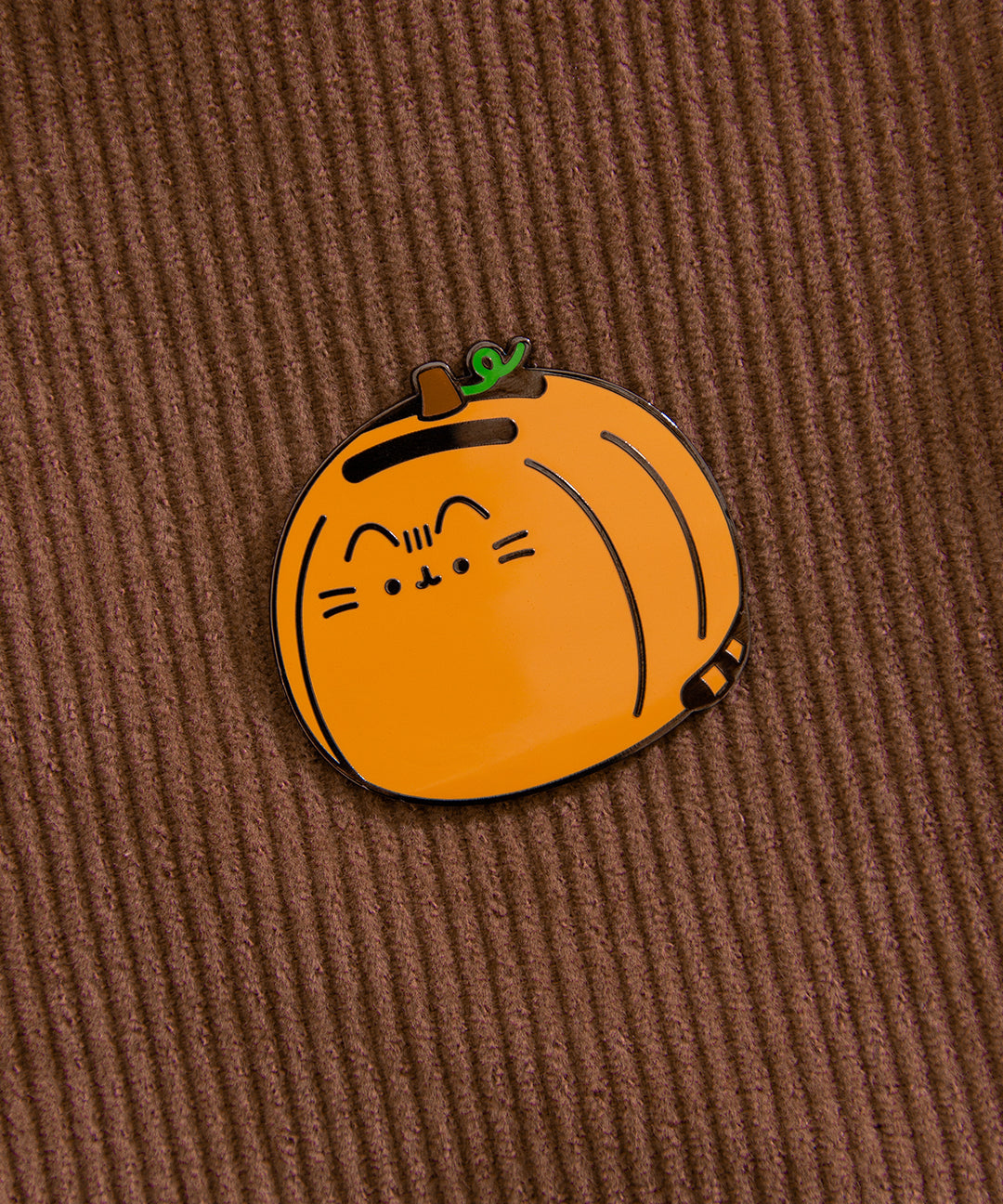 Halloween pumpkin pin attached to brown corduroy. The close-up view of the show the black outline of the pin and Pusheen face details on the orange pin.