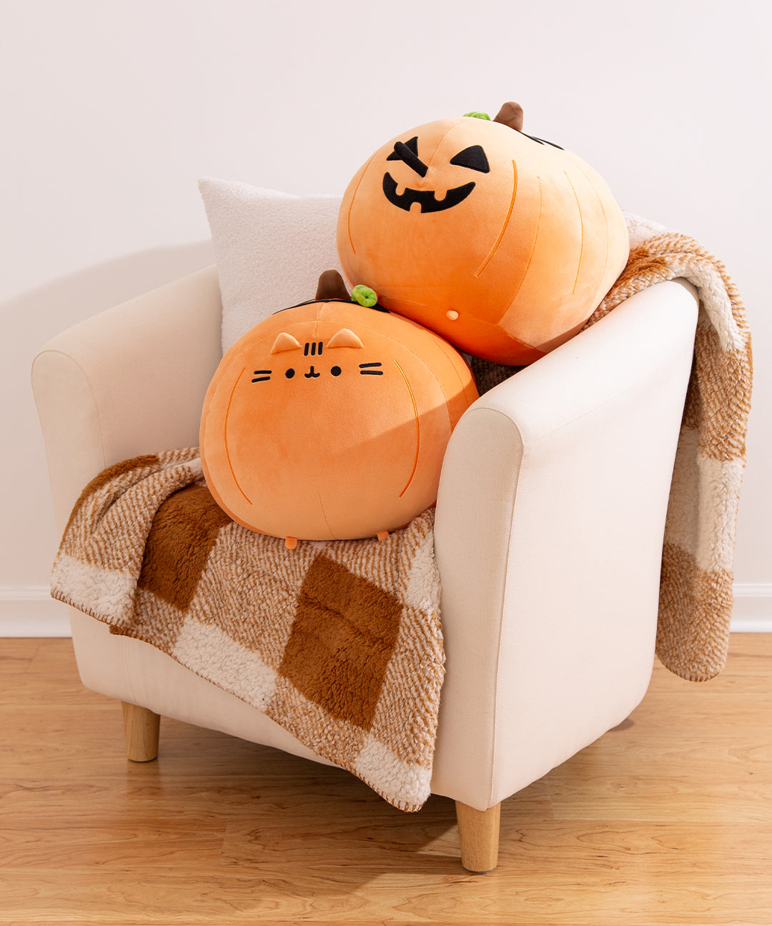 Two Big Mochi Pumpkin Squisheen Plush sit stacked on a chair. The front of the plush shows Pusheen's face and 3D triangle ears. The back of the plush shows a jack-o'-lantern face with a 3D tail as the nose.