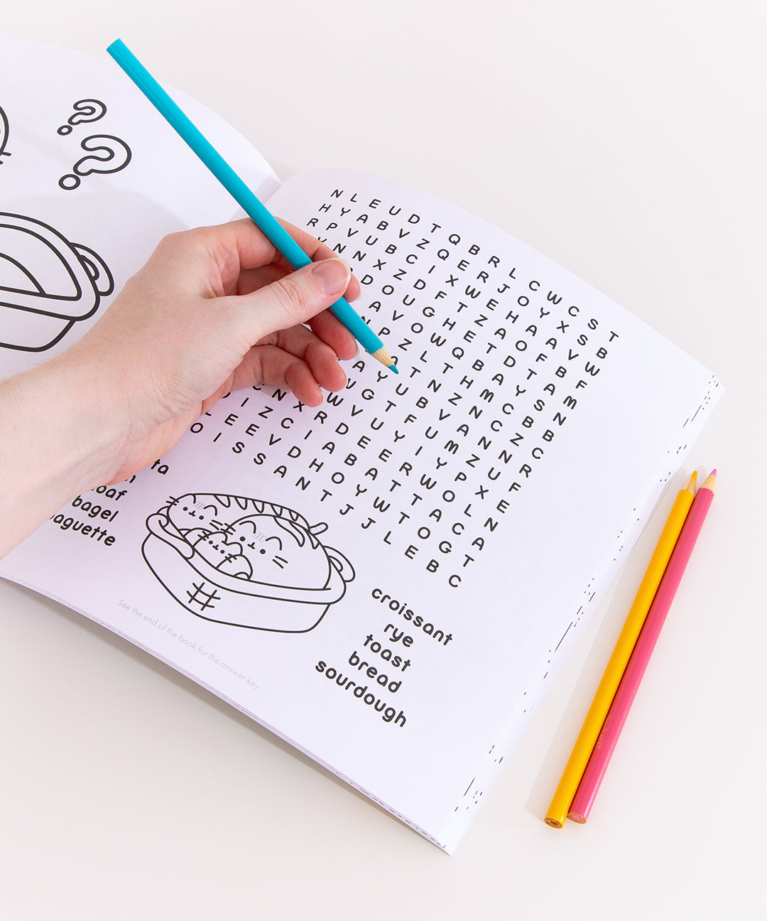Model holds a blue colored pencil above a crossword puzzle page of the Pusheen activity book. The crossword puzzle has many bread-themed words including croissant. Sourdough, and toast. Under the crossword puzzle is a graphic of a breadbasket holding Pusheen taking the form of a baguette, croissant, and loaf of bread.  