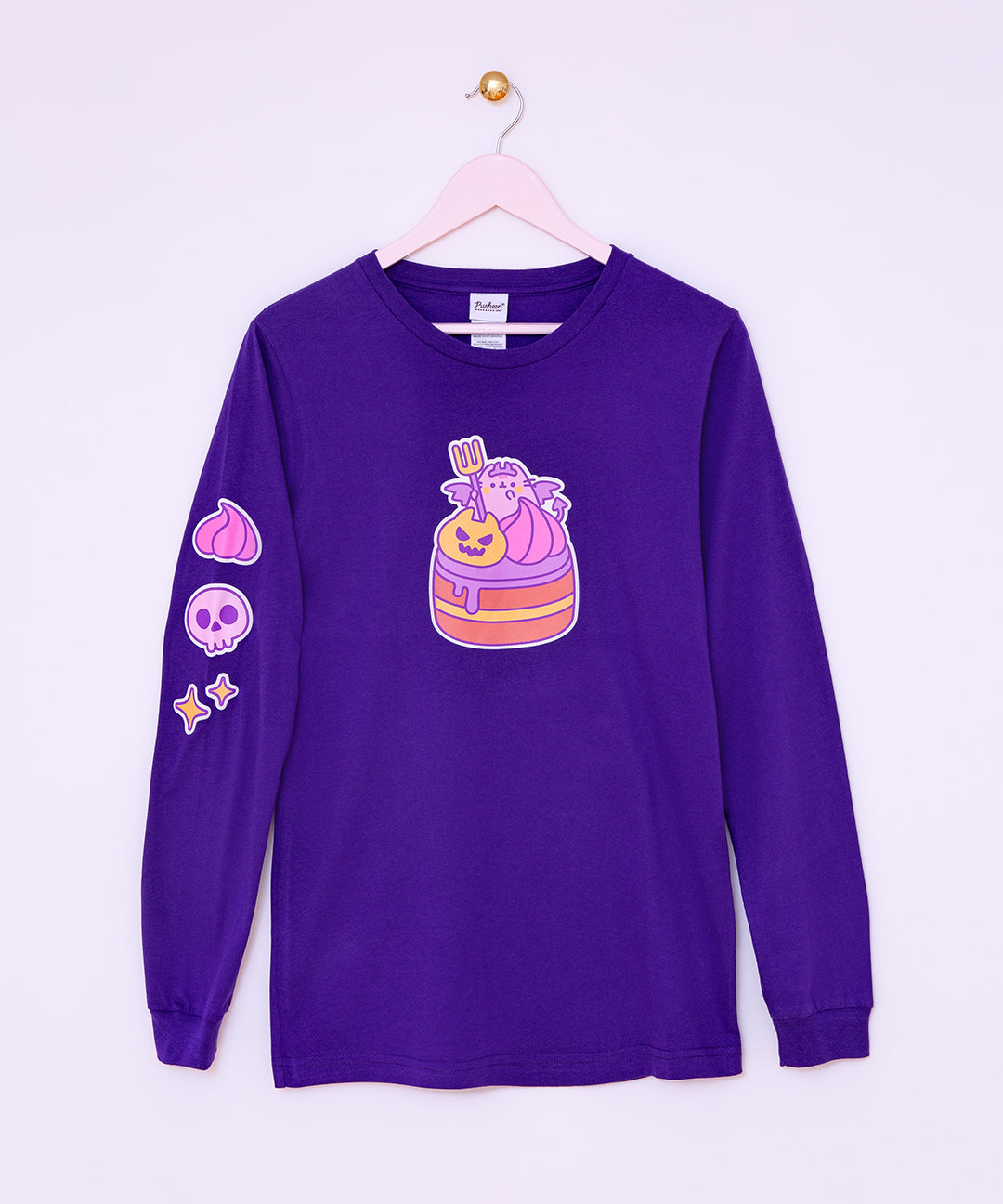 The Devil’s Food Cake Pusheen Long-Sleeve Unisex Tee hangs on a light pink hanger in front of a light purple wall. The dark purple tee shirt features Pusheen the Cat as a flying devil holding a pitch fork while sitting atop a layered devil’s food cake. The large, center front graphic is yellow, pink, purple, orange, and brown is accompanied by a pink whipped dollop, purple skull, and yellow sparkles graphic down the wearer’s right sleeve. 