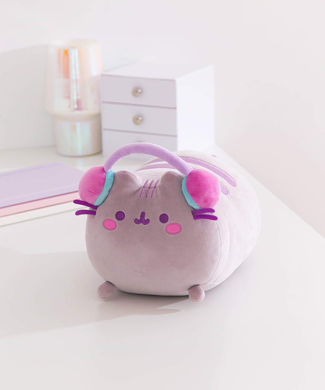 Front view of the Gamer Pusheen Log Squisheen Plush. The long cat plush toy wears a set of purple, pink, and teal headphones that extend over the top of her head.