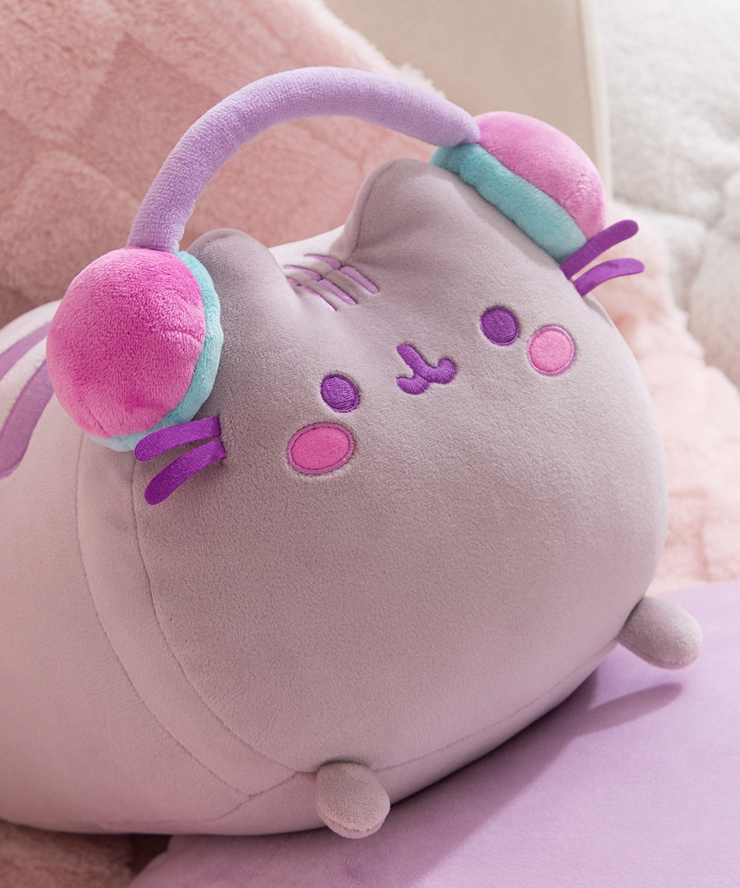 Close-up of the long gaming log plush toy. The grey cat has purple facial features embroidered on her face including eyes and mouth. Pusheen's classic three head stripes are embroidered between her two triangle shaped ears.