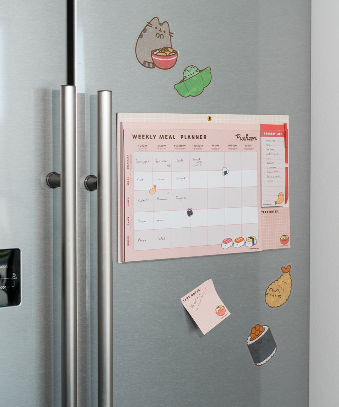 The Pusheen Magnetic Meal Planner is attached to a stainless steel refrigerator. A sticky note is stuck below the pad with a note written on the page.