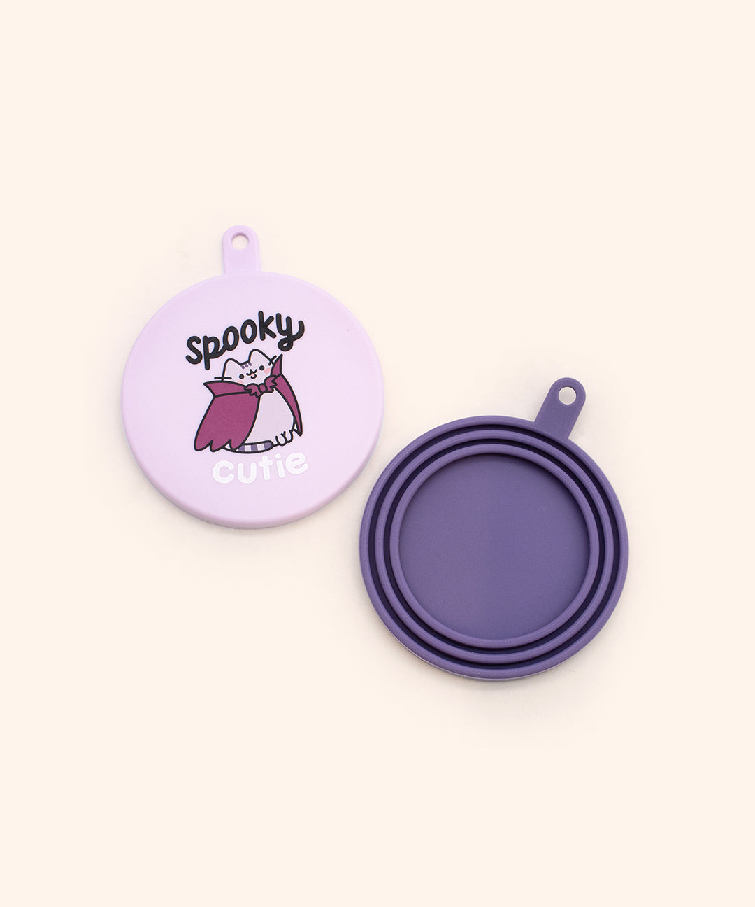 Close-up of the purple pet food can covers where one is dark purple and one is light purple. The two silicone can covers have graphics printed on the top of spooky Pusheen. The dark purple can cover back is shown to showcase that the cover can fit multiple can sizes.