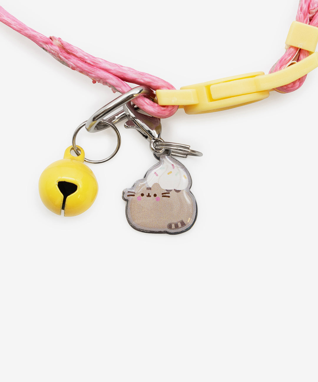 Close-up of the included bell and collar charm on the pink pet collar. Both are attached to the collar using metal fasteners. The bell is yellow and the charm has a graphic of Pusheen with a dollop of whipped cream and sprinkles on her back.
