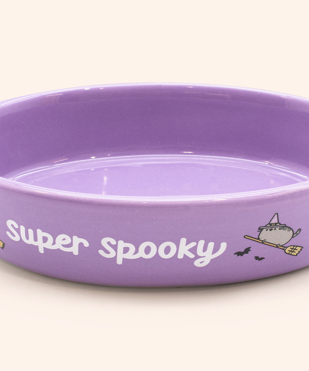 The Pusheen Pet Purple Halloween Bowl has a Pusheen print on the outer edge of the purple ceramic pet bowl. The print features a repeating pattern of Pusheen wearing a Witch hat and riding a broom with the phrase "super spooky"