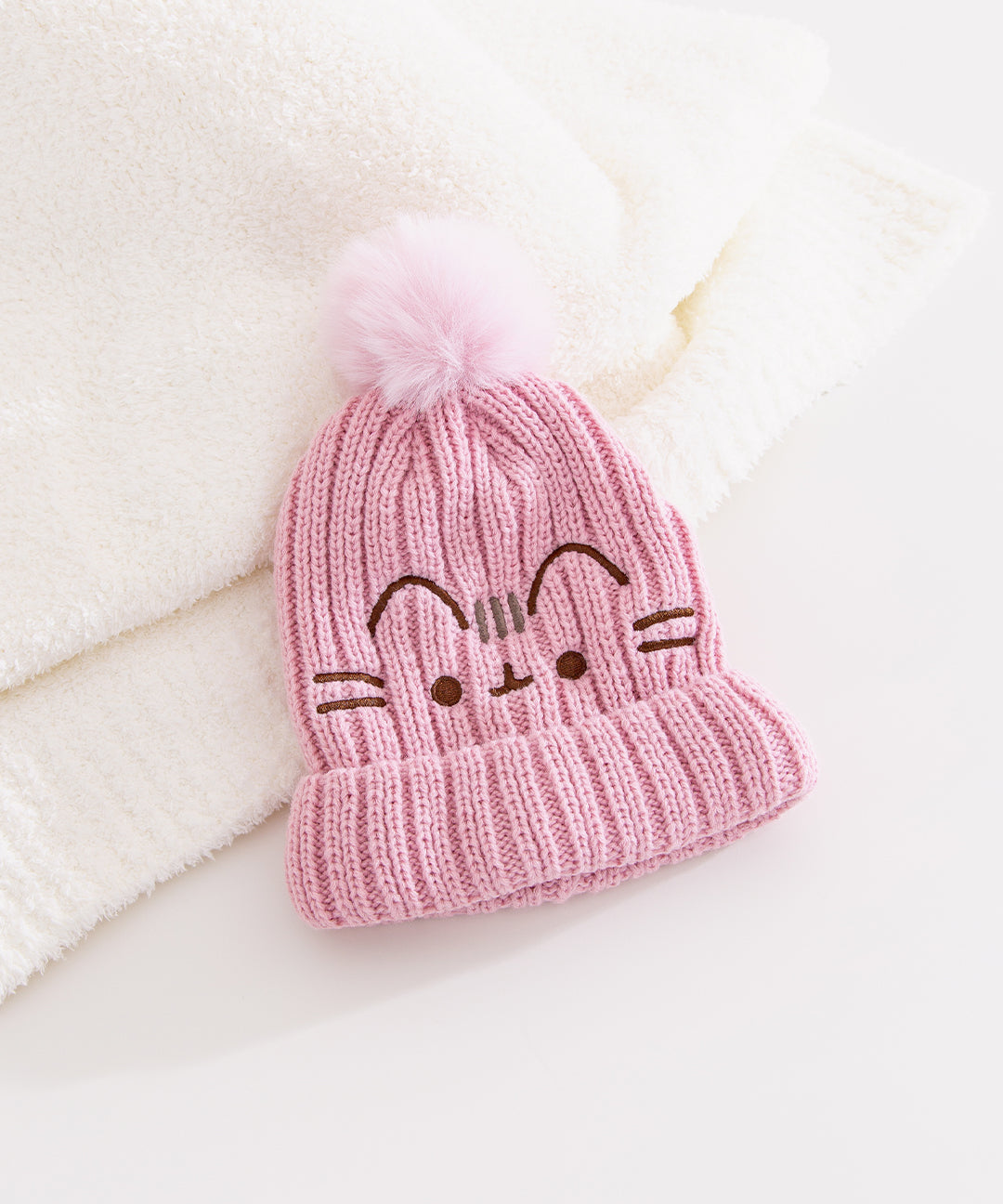 Pink knitted Pusheen beanie with a pink pom-pom is propped up against a white blanket on a white background.