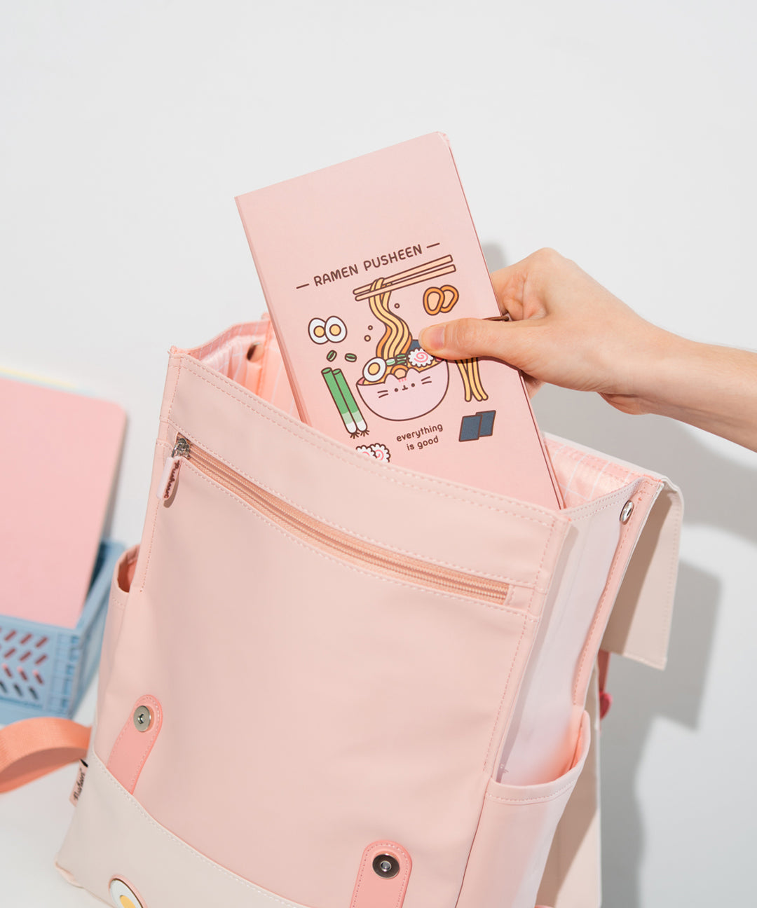 Ramen-themed backpack open to shown interior while model places a notebook inside. The front exterior zip pocket and one side pocket can be seen.