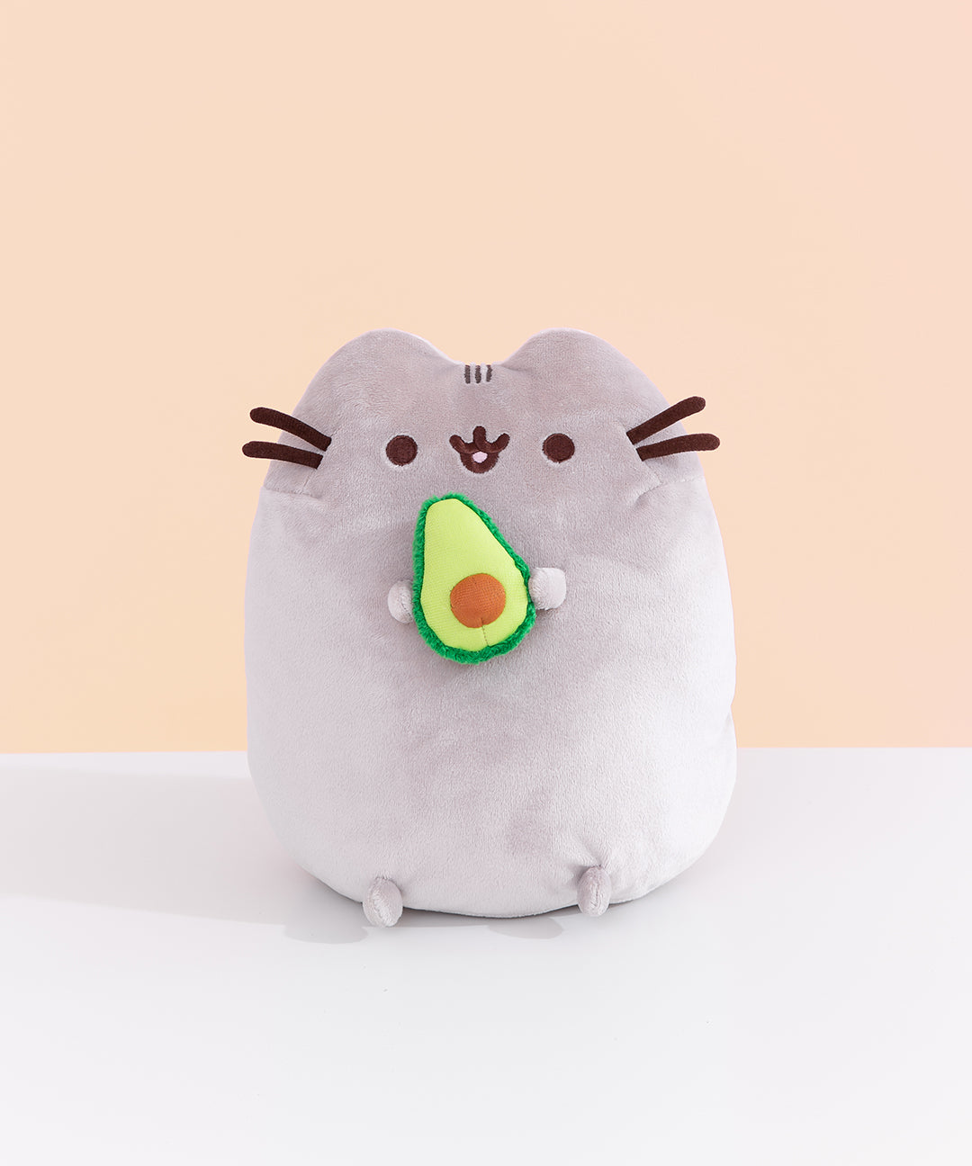 Front view of Pusheen Avocado Plush. Pusheen the Cat is shown with her classic brown eyes, mouth, and whiskers. Pusheen holds a cut-in-half, two-toned green avocado with a light brown pit.