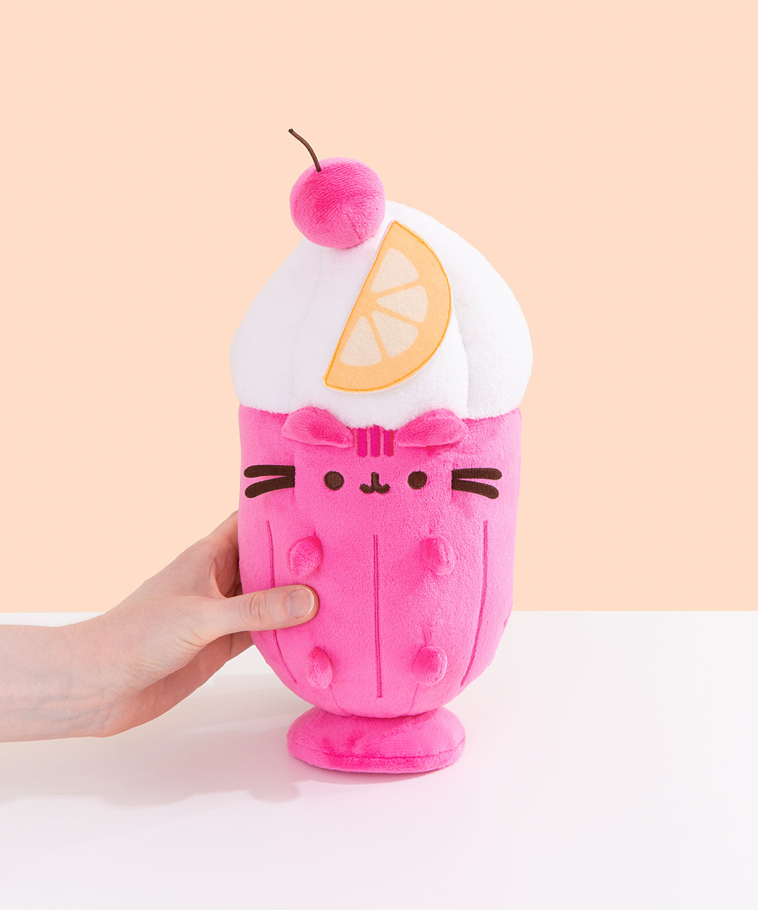 Model holds the side of the Parfait Pusheen Plush. The hot pink body is accompanied by vertical pink lines to imitate a classic glass container. Pusheen's head stripes, eyes, smiling, mouth and whiskers are embroidered on the front of the plush while her hot pink ears and paws extend off the plush body.   