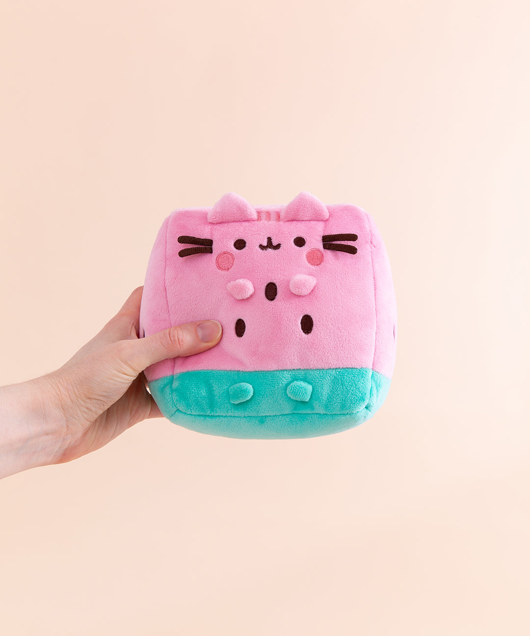 Model holding side of the Pusheen Fruits Watermelon Plush. Pusheen the Cat takes the form of a juicy pink and mint green watermelon slice. The triangle shaped plush body has embroidered brown cylinder shapes to represent watermelon seeds. On the front of the plush are 3D versions of Pusheen’s ears and four paws.  