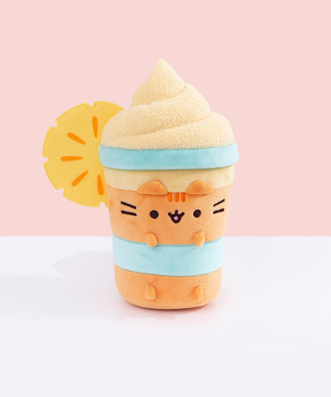 Front view of the Pusheen Fruits Pineapple Float Plush. Pusheen the Cat takes the form a delicious tropical drink served with whipped topping and a pineapple slice. The whipped topping is light orange and has a textured swirl extending off the cylinder-shaped body.