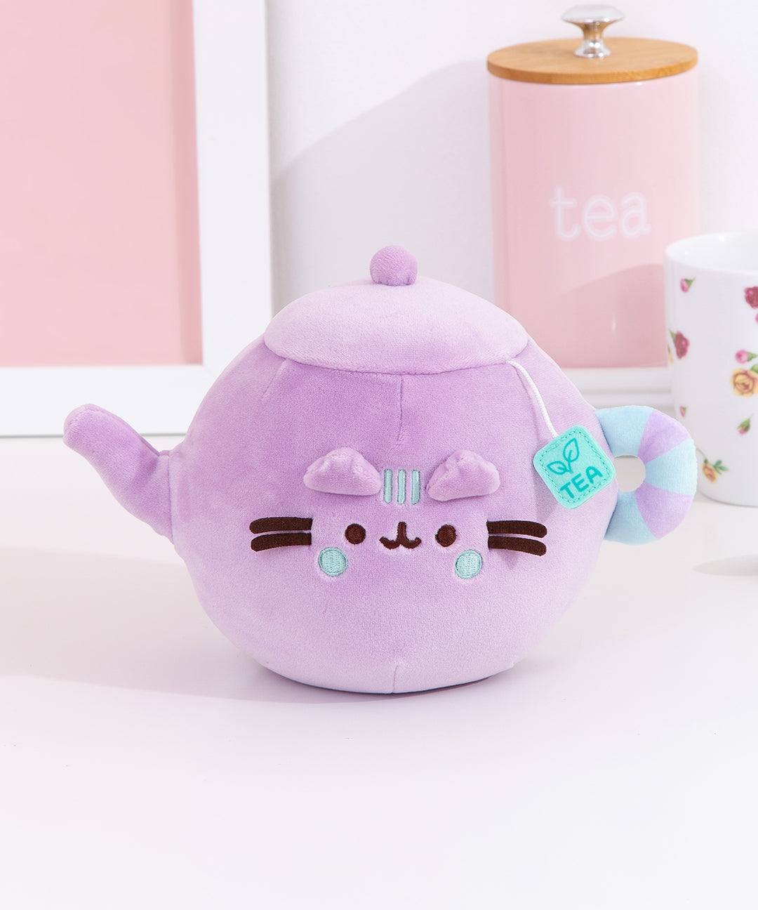 Front view of the Pusheen's Kitchen Teapot Squisheen Plush. The lilac colored plush shows Pusheen the Cat taking the form of a classic tea kettle with a tea bag label coming out of the lid.