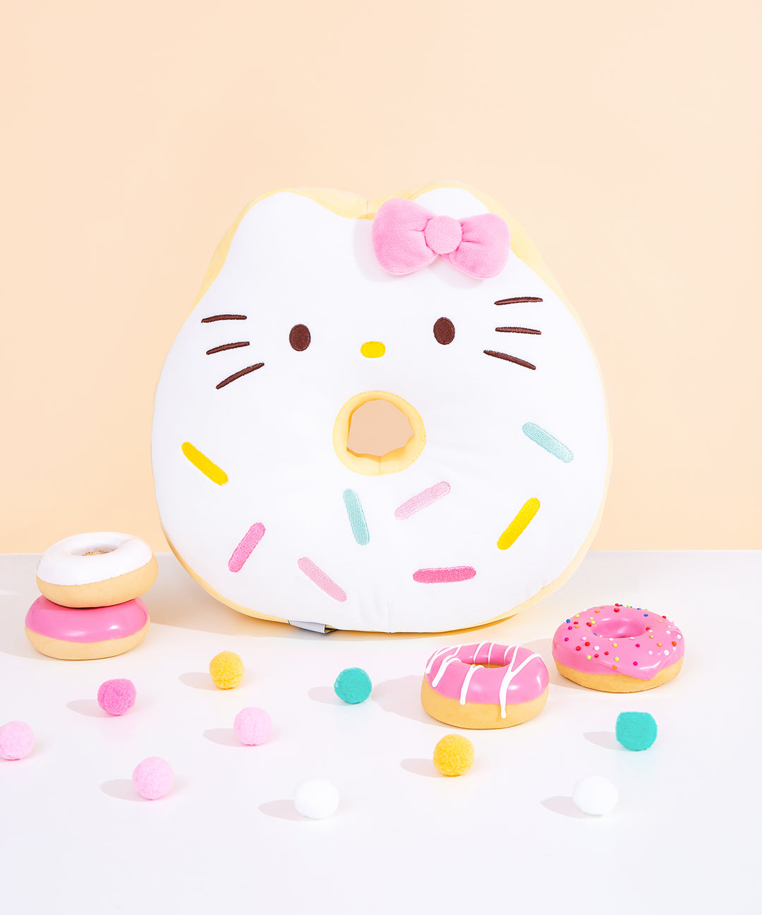 Front view of Hello Kitty side of Hello Kitty x Pusheen Donut Plush. The double-sided donut inspired plush showcases Hello Kitty as a white glazed donut with multicolor blue, pink, and yellow sprinkles. Hello Kitty is accompanied by her classic pink bow. Plush is surrounded by pink and white donut figurines and multicolor poms.  