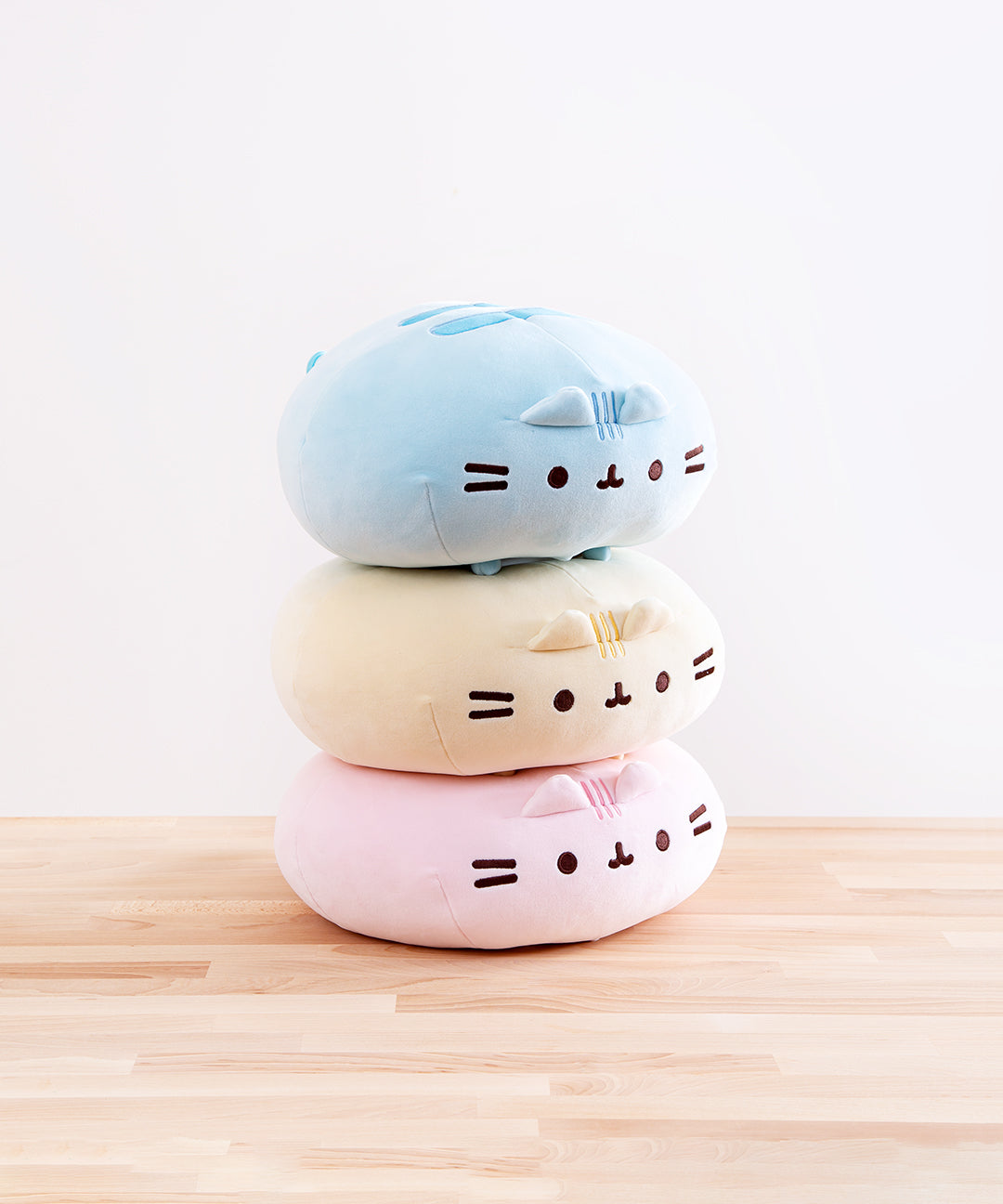 The three Round Squisheen Plush stacked on top of one another, sitting on top a wooden floor in front of a white background. The pink plush is at the bottom, the yellow in the middle, and blue on the top. The pink and yellow plush are squished down enough to look as if they have no feet, but the blue round Pusheen still has her feet.