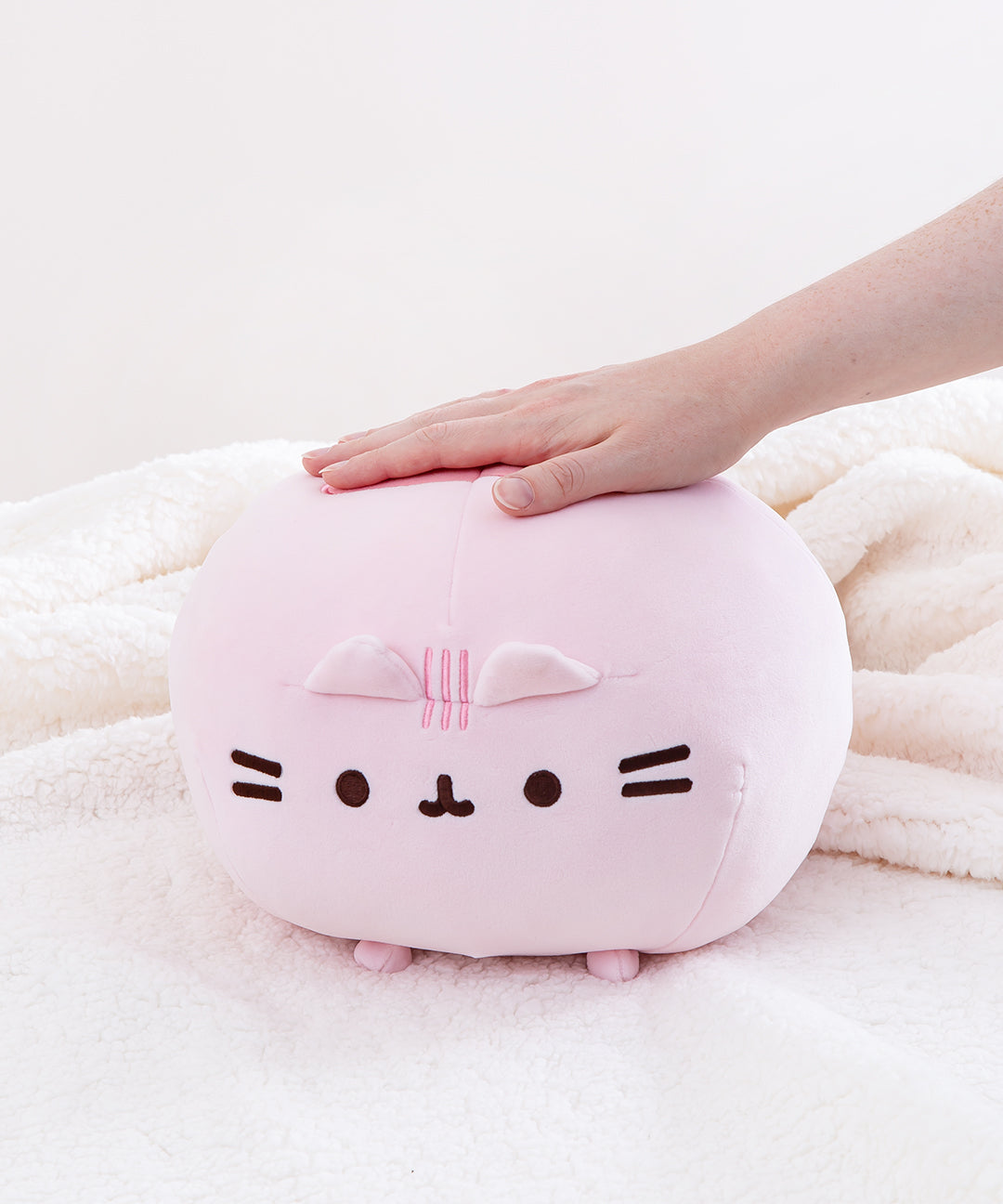 Front view of the Pink Round Squisheen sitting on top a fluffy white blanket, a model’s hand sticking out from the right to rest on top of the plush.