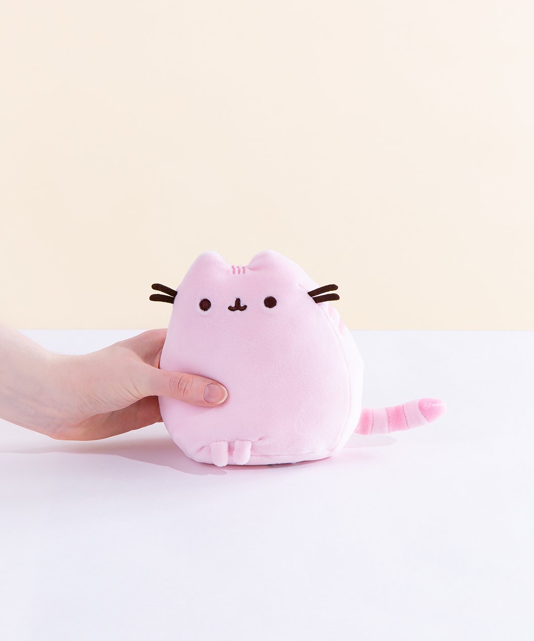 A model’s hand holding the pink Mini Squisheen Plush in front of a white and pale yellow background. The pink plush has dark pink stripes.