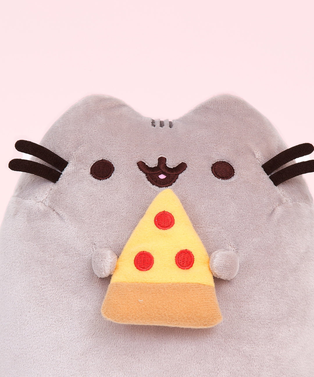 Closeup view of Pizza Pusheen Plush. In her paws, Pusheen holds a yellow, red, and light brown slice of pizza representing pepperoni pizza. She is shown with her classic black and brown nose, eyes, and whiskers.