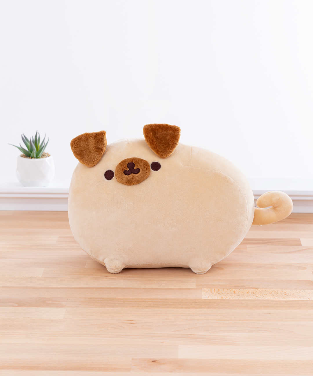 Light brown round dog with a circular brown snout, droopy brown ears that look like triangles, and a curly tail. Pugsheen has the same embroidered dark brown dot eyes and cat mouth as Pusheen the Cat does, and is standing in the same pose as the default Pusheen pose. The plush is in front of a white wall on top of a wooden floor, and a potted succulent can be seen to the left of the plush..