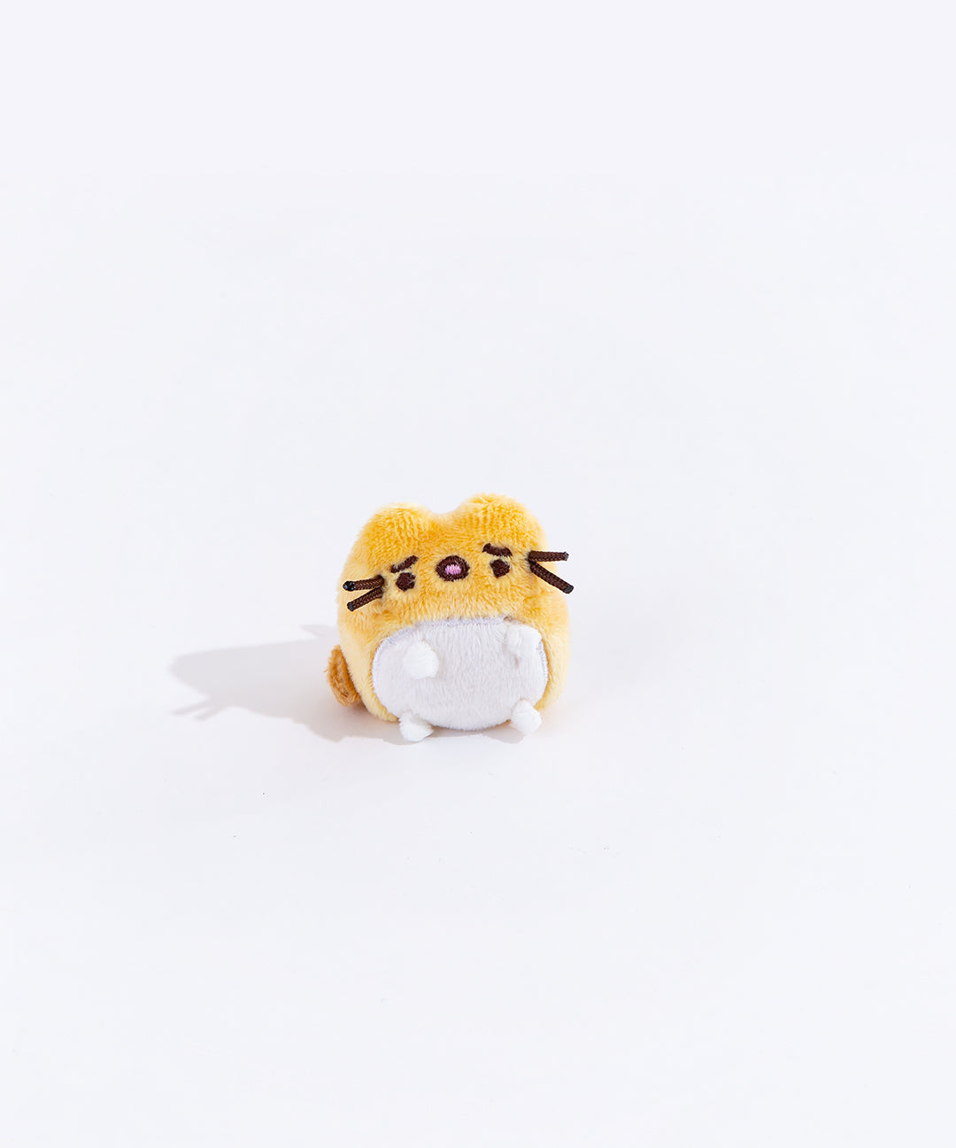 Full view of the mini worried yellow cat plush from the collectors set. The cat is a golden yellow, with a dark orange tail on the left side. Their belly has a white circle, which encompasses their feet. The cat has worried eyebrows embroidered into it, and their mouth is open.