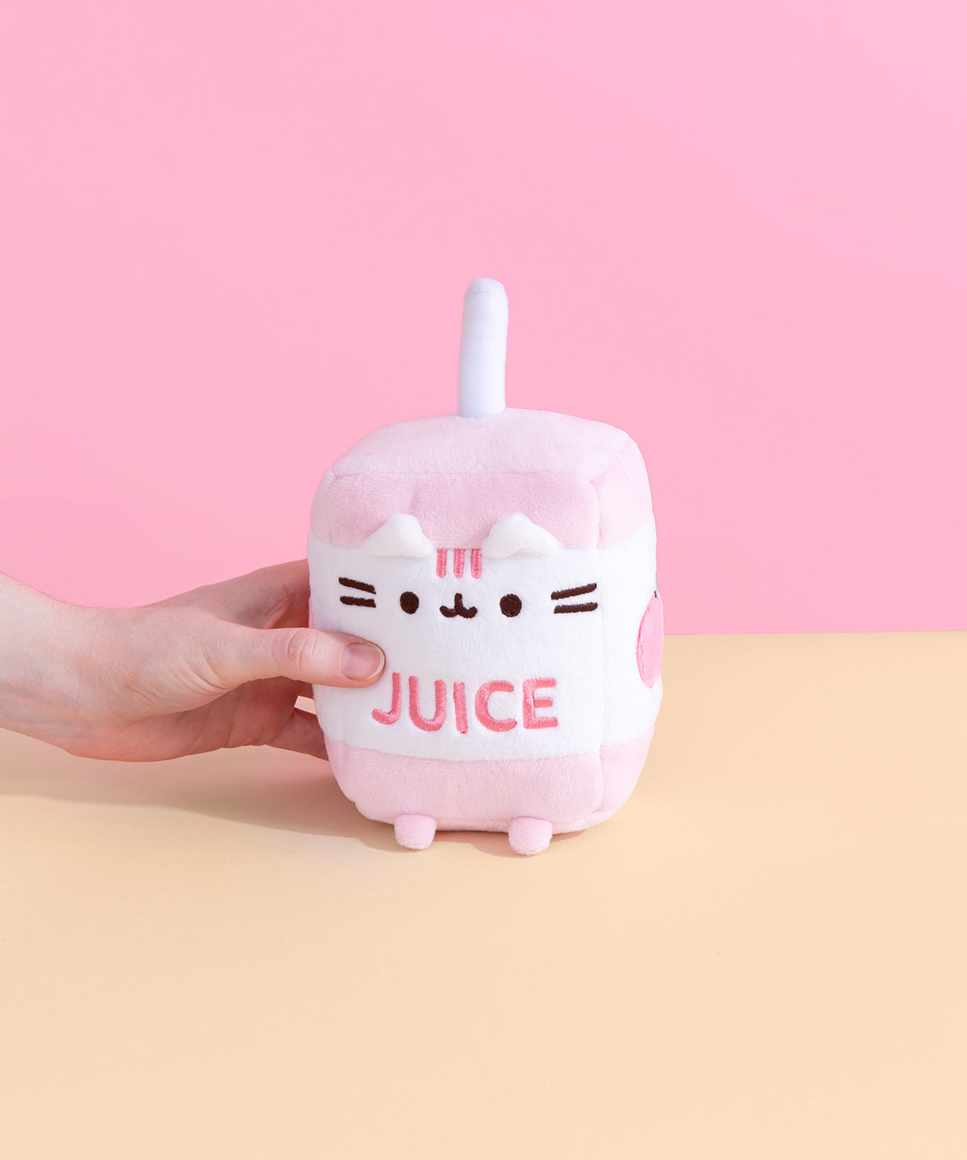 A model’s hand lightly squeezes the Juice Box Sips Plush in front of a pink and yellow background.  Pusheen is in the shape of a juice box, with a plush straw sticking out on top. The top and bottom of the box are light pink, and the middle is white, with an embroidery of Pusheen’s face, the word ‘juice’ in pink, and a partially visible graphic on the right side. There are white plush ears sticking out under the rim of the box, and pink plush feet sticking out from the bottom. 