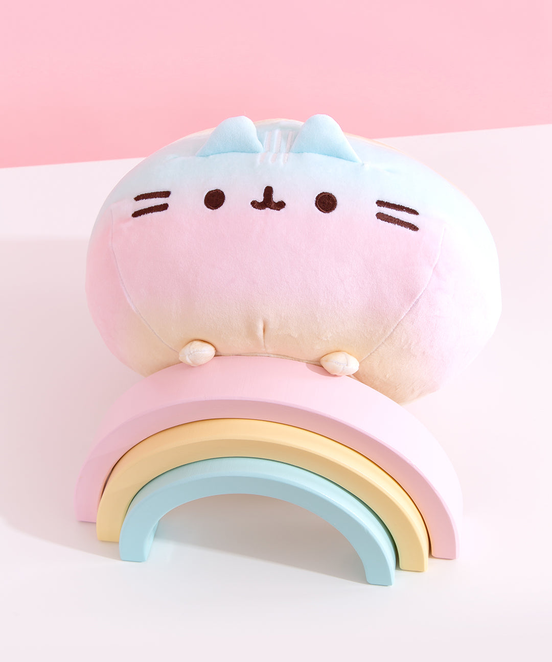 Rainbow Pusheen plush is angled on top of a pink, yellow, and blue pastel wooden rainbow. The plush’s paw details are yellow.  