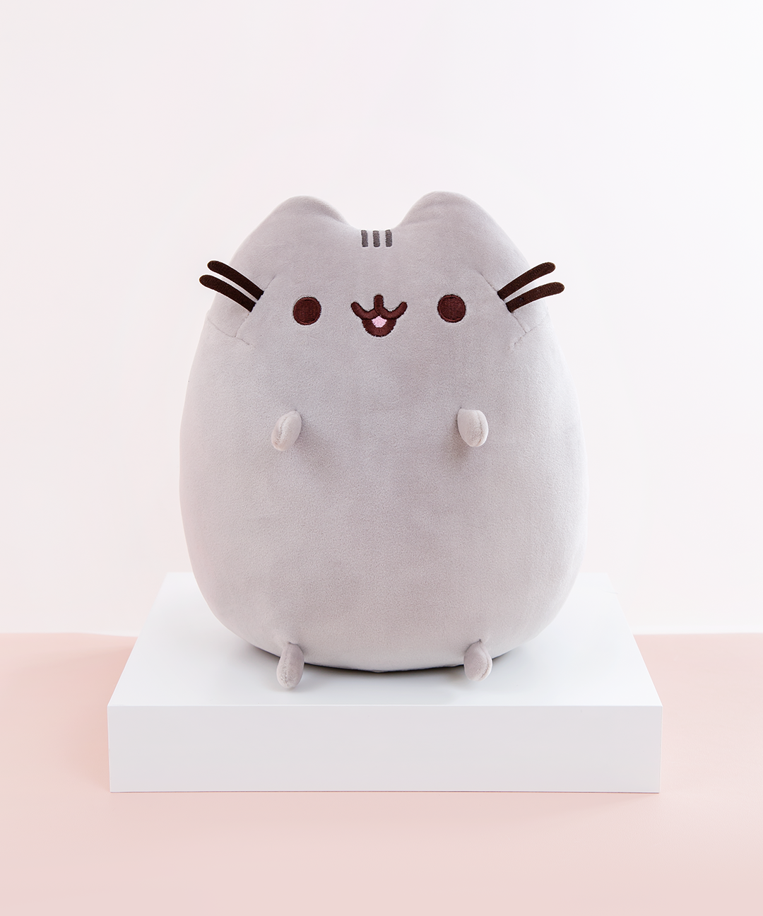 Pusheen sitting upright, with two nub paws up in the air and two nub paws at the bottom facing forward. Pusheen’s eyes and slightly open mouth are embroidered onto her face, and she has two felt whiskers poking out from each side of her face.  The medium sitting squisheen is on top of a white square podium, on top of a peach floor in front of a white background.