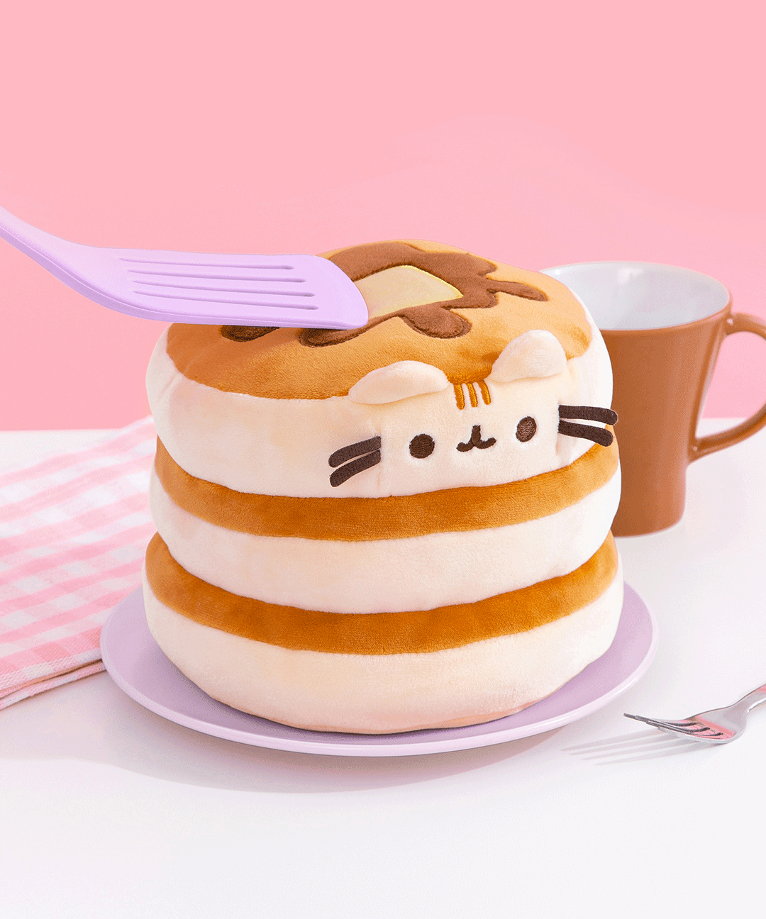 Purple spatula presses down on pancake squisheen stack to show how soft and squishy the plush is. Plush is placed on a purple plate and surrounded by a brown mug, white and pink dish towel, and silver fork.